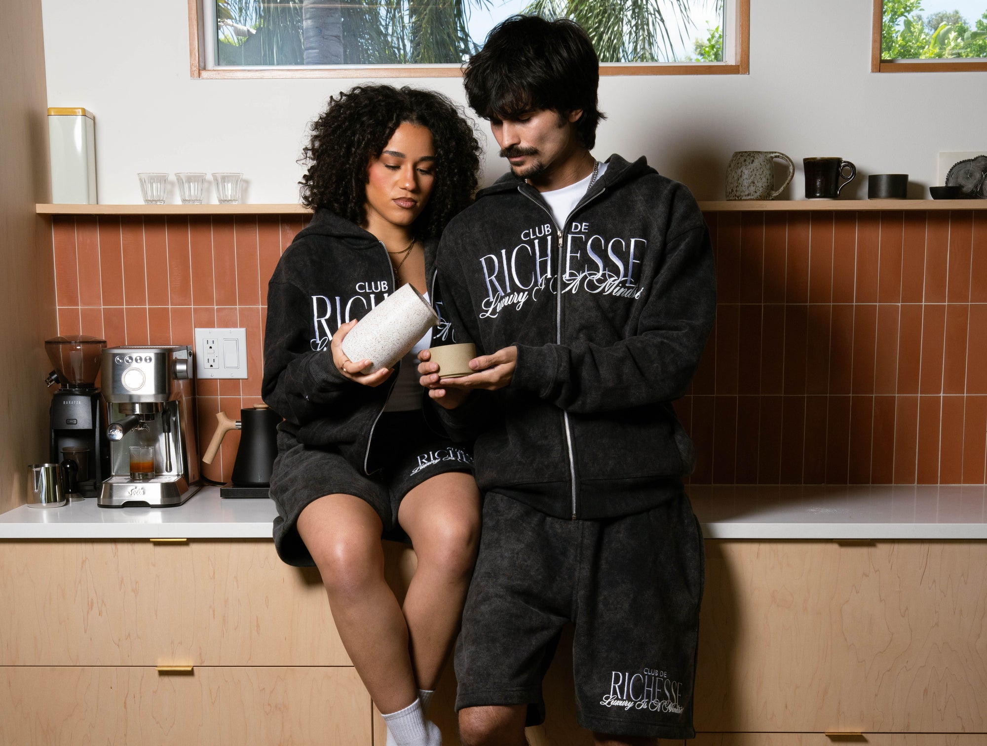 Two people wearing matching black tracksuits with 'Club Richesse' branding in a kitchen.